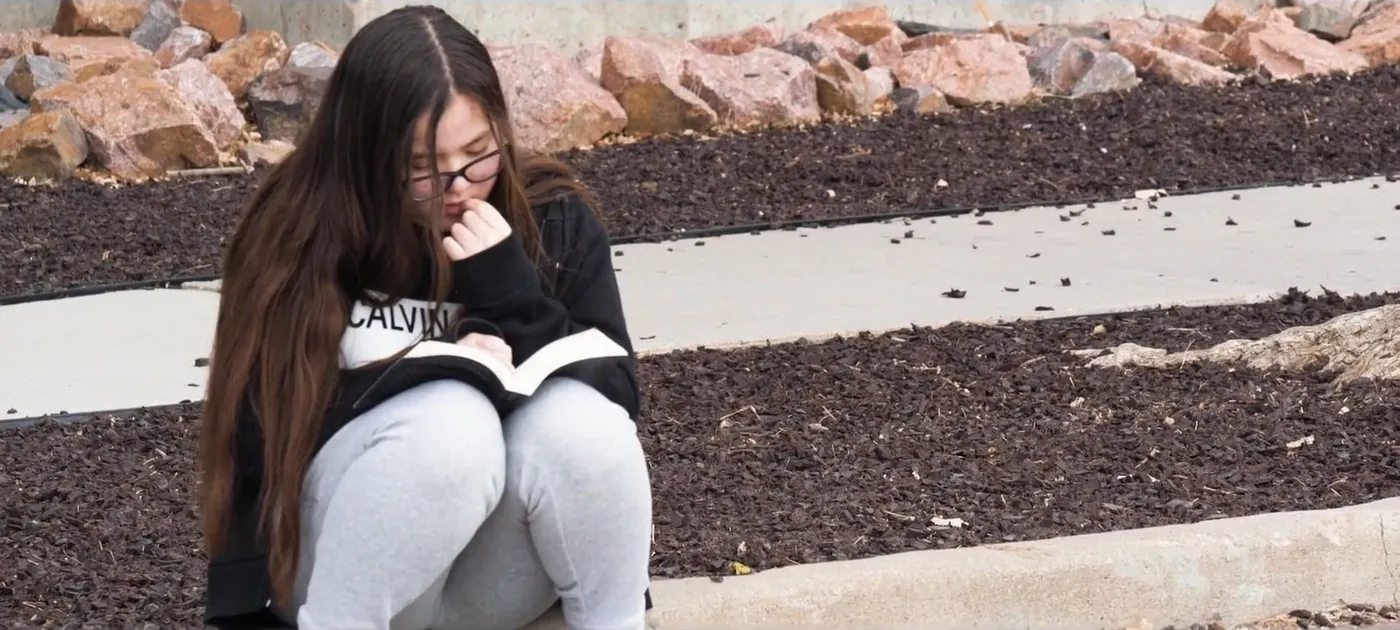 A girl with long dark hair and glasses reads a book, sitting on a kerb outdoors.