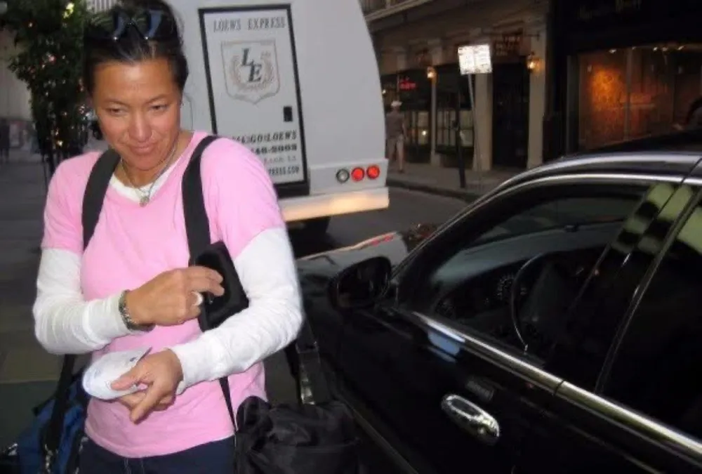 Scilla in a pink T-shirt with a backpack, on a city street beside a black car and a hotel shuttle.