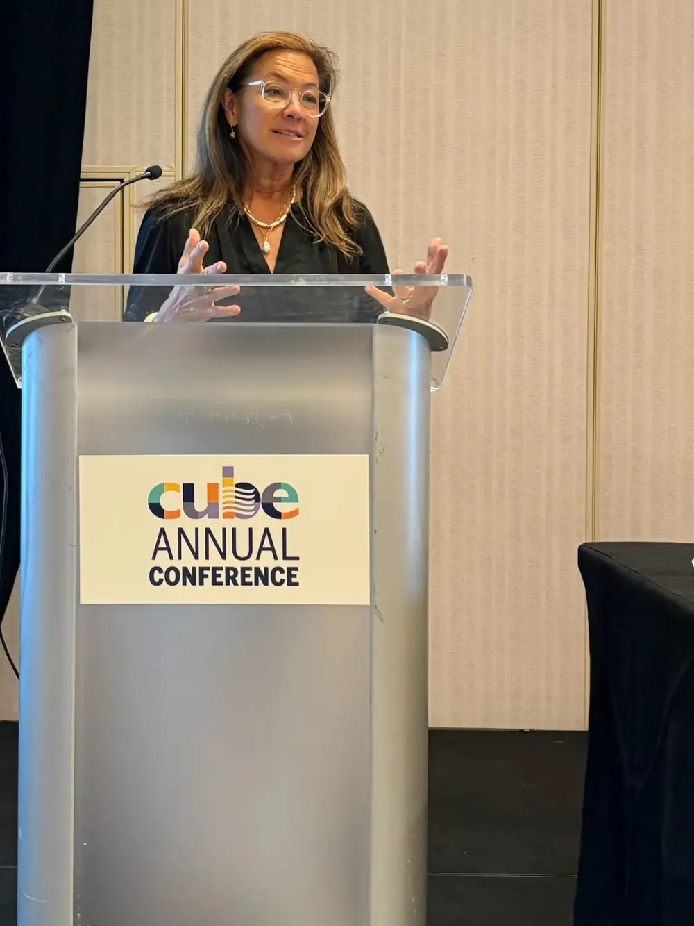 Scilla speaking at a clear podium marked CUBE Annual Conference, glasses on, hands open.