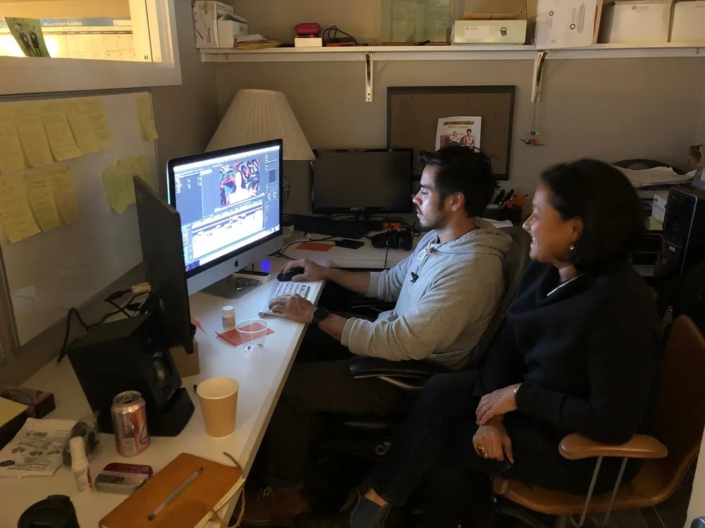 Scilla and a young man at an editing desk, watching footage on a big monitor, sticky notes covering the wall beside them.