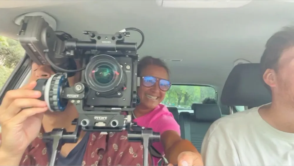 Scilla in sunglasses and a pink top, laughing behind a cinema camera rig in the back of a car, a young man beside her.