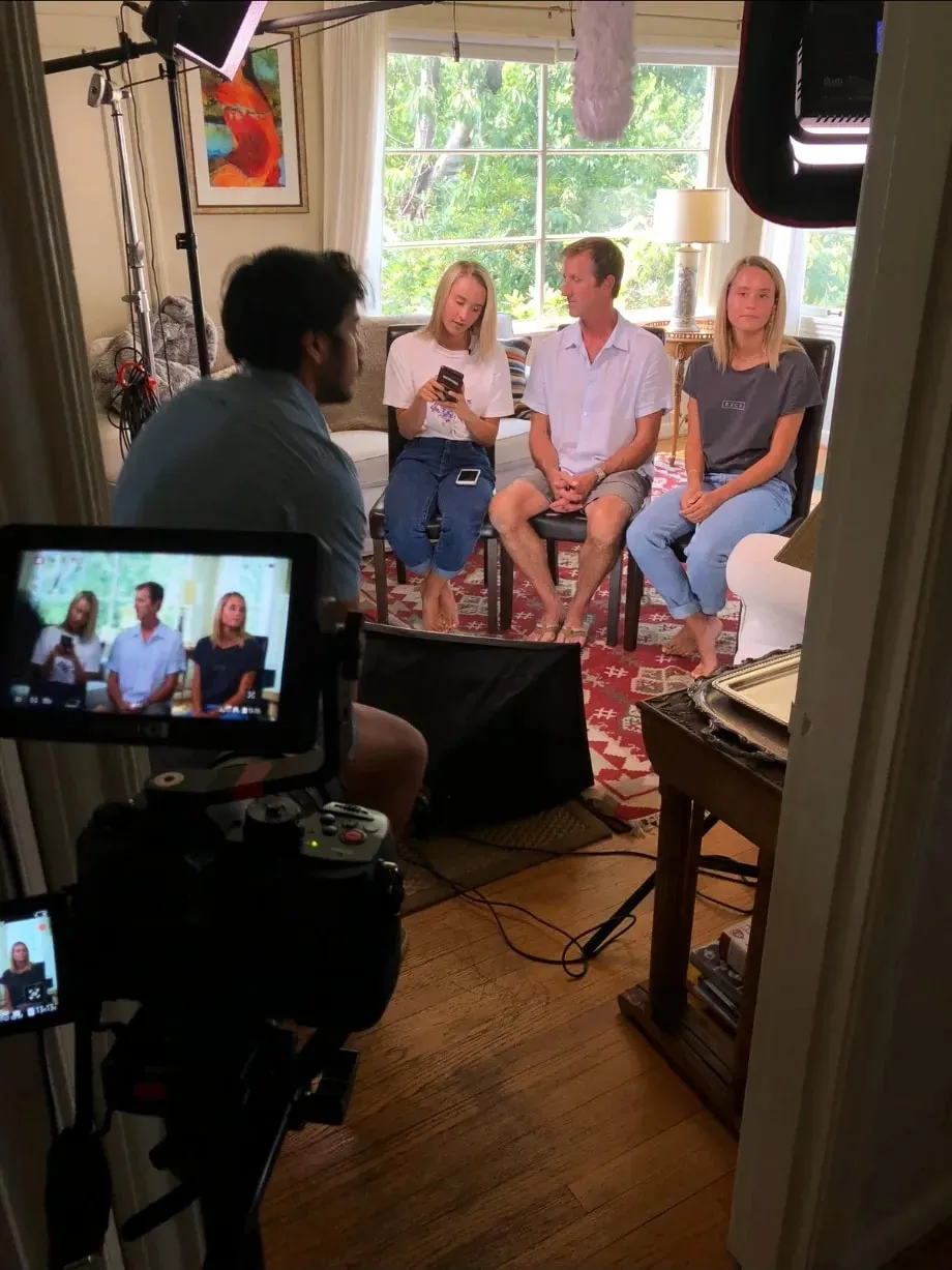 Scilla’s son Ian, the director of photography, filming her twin daughters, Arin and Ally, and her husband in a sunlit living room, seated side by side, with the camera monitor in the foreground.