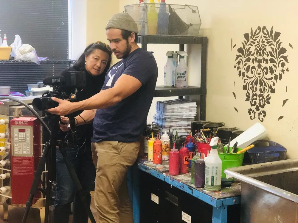 Scilla and a young man in a beanie look together at a camera’s screen on a tripod, in a busy art studio.