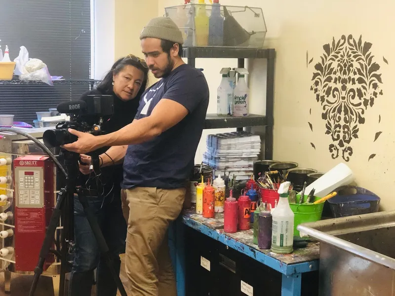 Scilla and a young man in a beanie look together at a camera’s screen on a tripod, in a busy art studio.