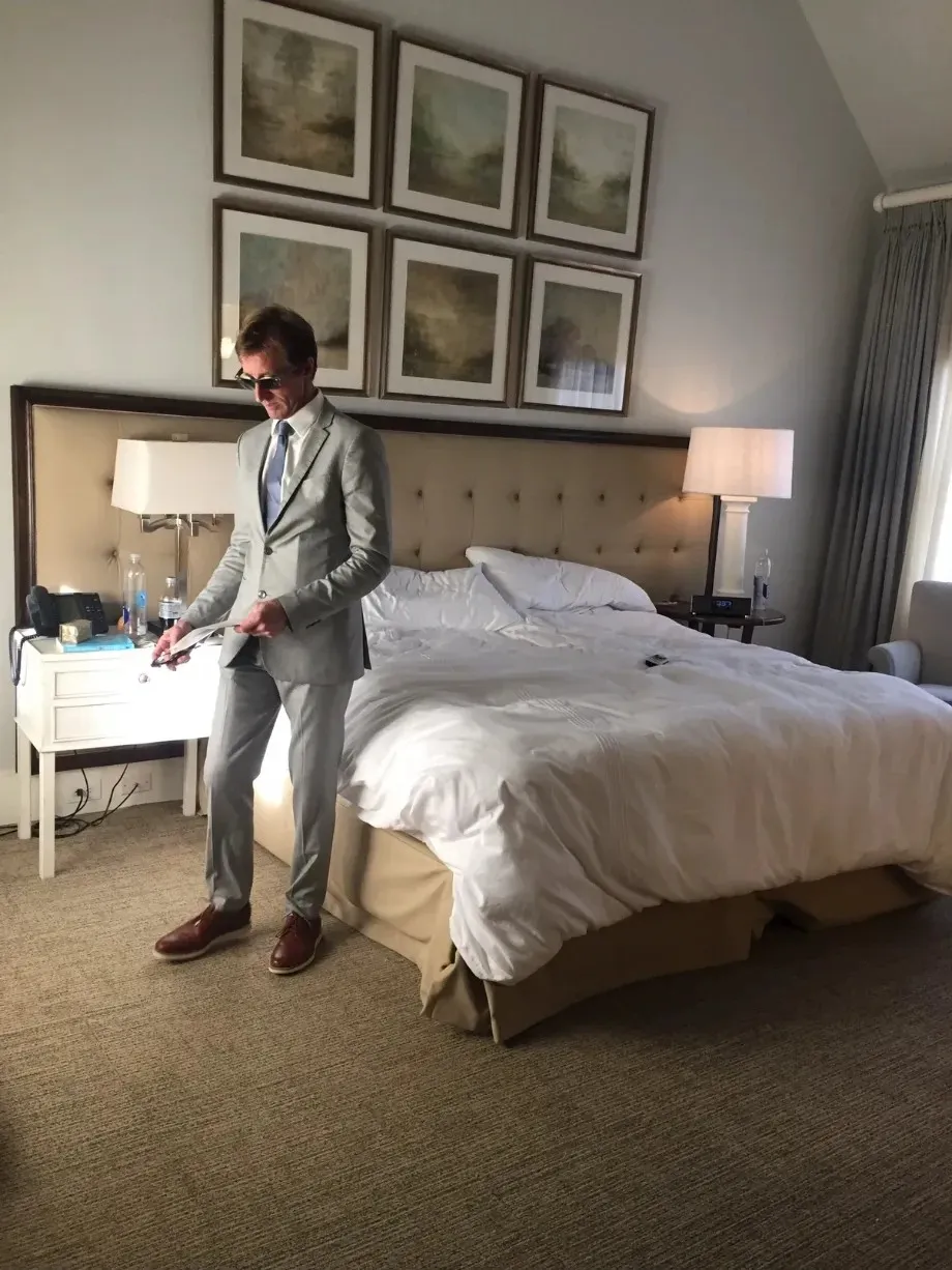 Scilla’s husband, Eric, in a light grey suit and sunglasses, reading notes in a hotel room on location.