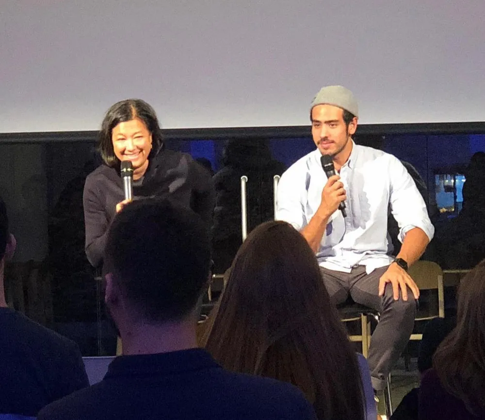 Scilla and Ian, each with a microphone, talking with an audience in front of a screen.
