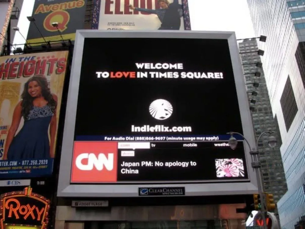 A giant Times Square billboard reading “Welcome to LOVE in Times Square! indieflix.com”, among theatre ads and skyscrapers.