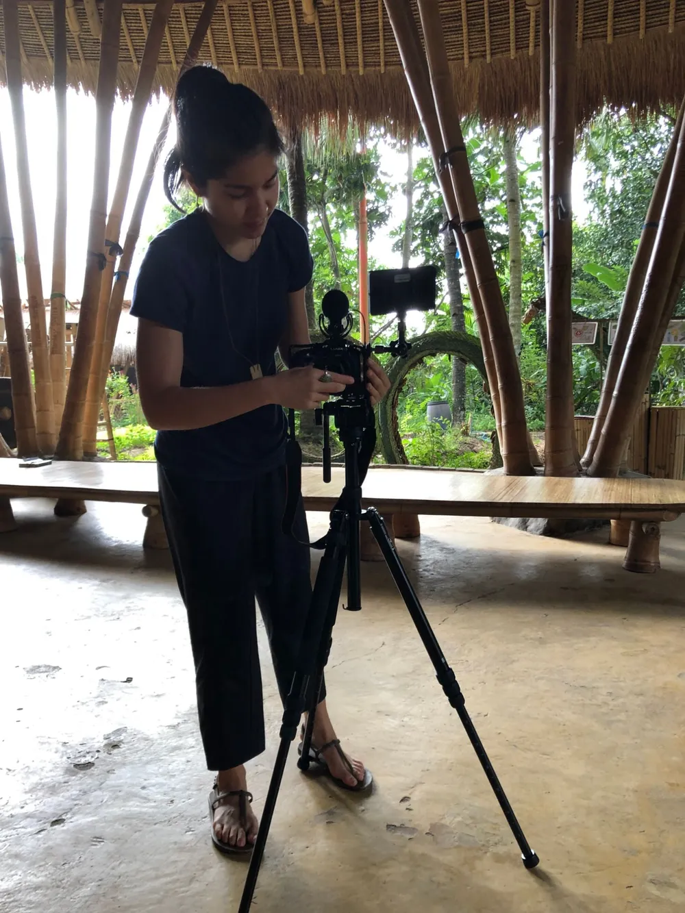 Scilla’s daughter Natalie setting up a camera on a tripod in an open bamboo building in Bali, jungle behind her.