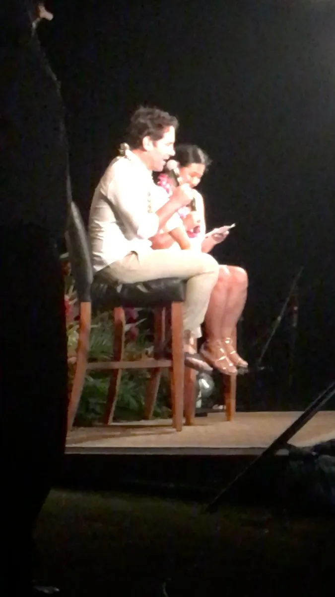Paul Rudd, wearing a lei, speaks into a microphone on stage beside Scilla, who reads questions from her phone.