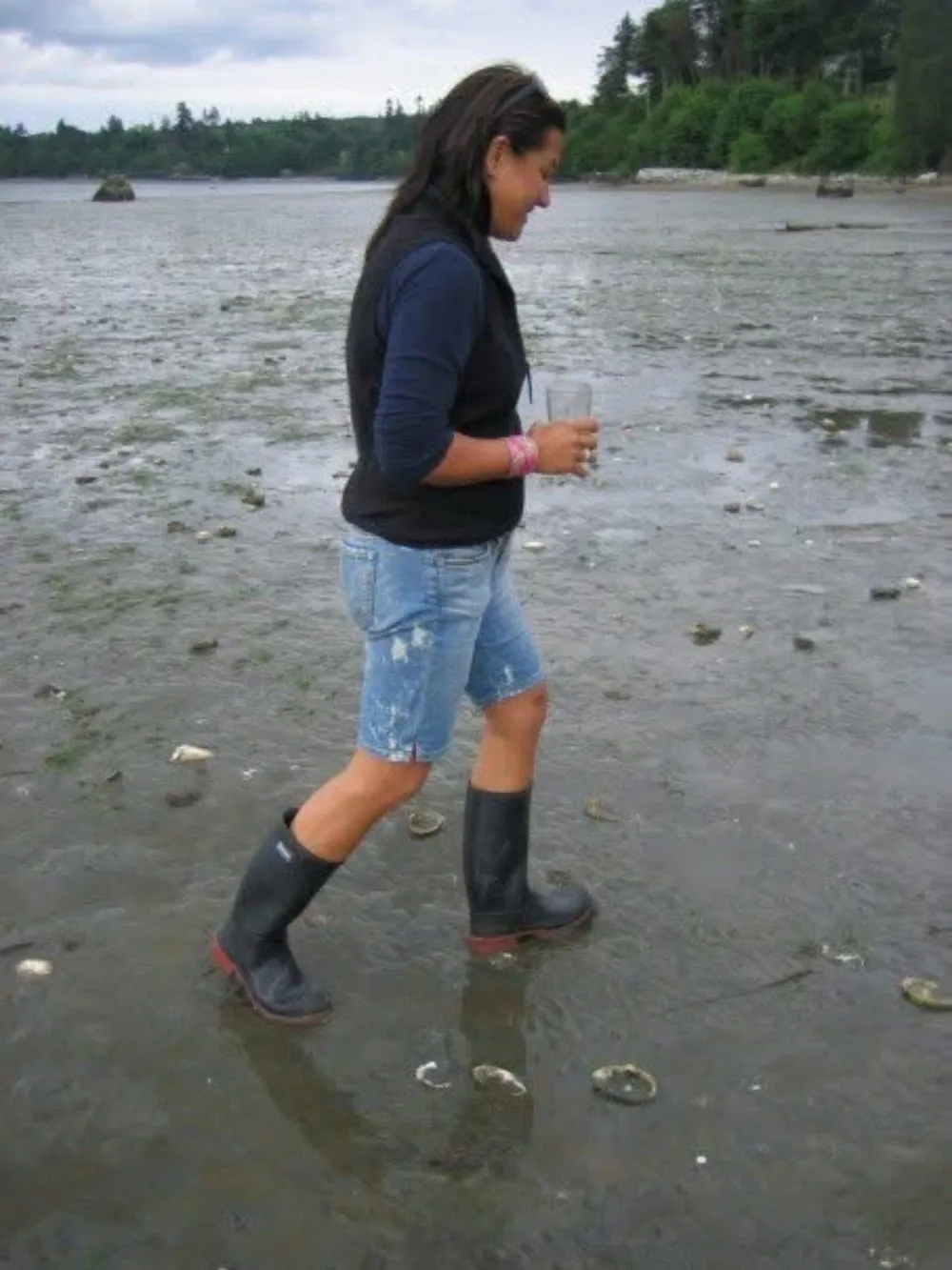 Scilla in rubber boots and denim shorts, walking across a tidal flat with forest and water behind her.