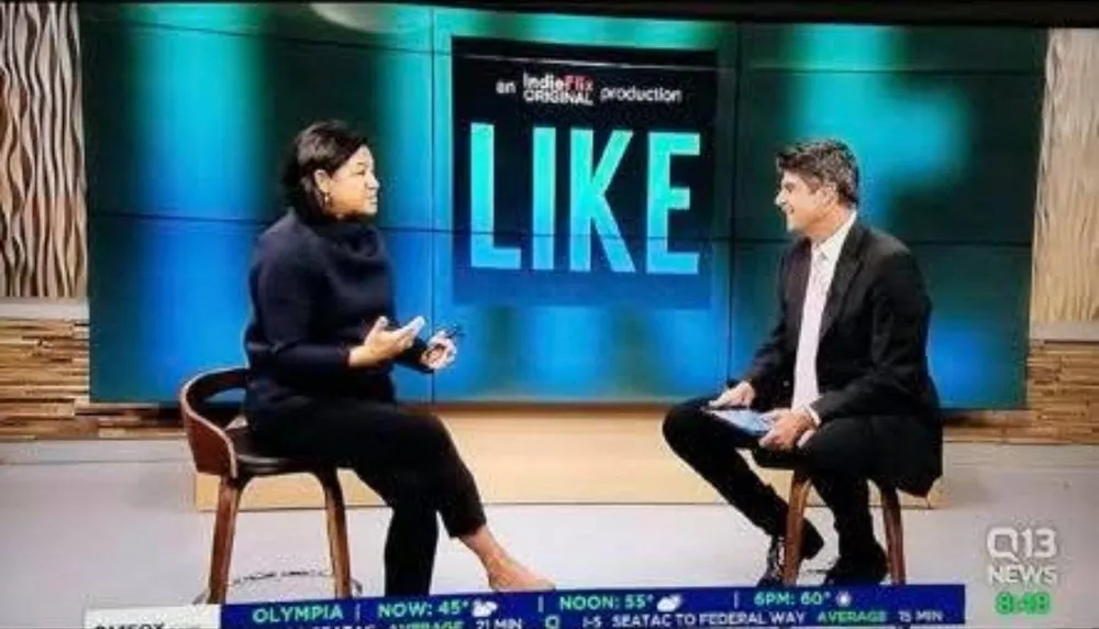 A television interview on Q13 News, two people seated in front of a screen showing the title LIKE.