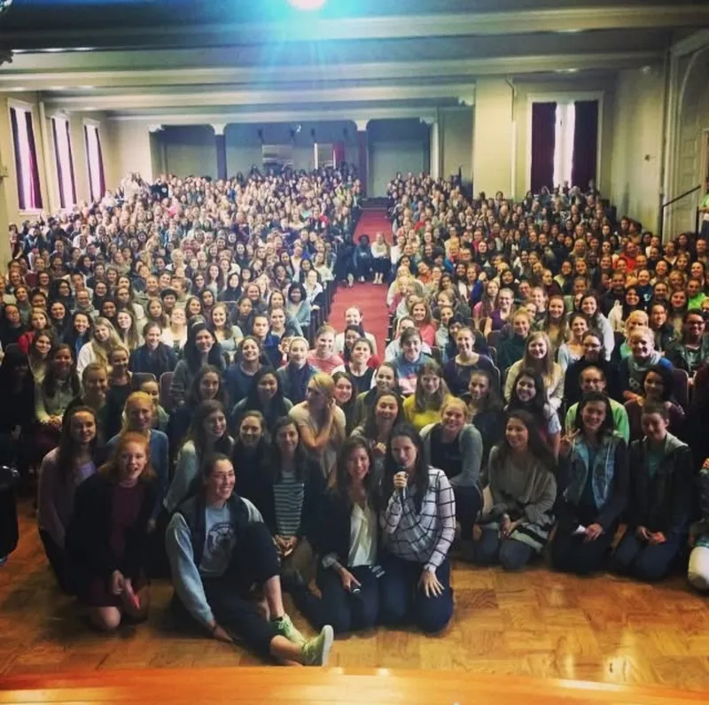A packed school hall of students, sitting on the floor at the front and filling every seat behind, smiling toward the camera.
