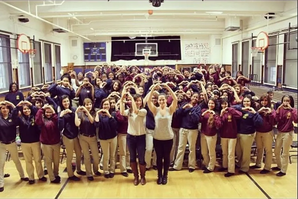 A gym full of students in school uniform making heart shapes with their hands, with two women at the front doing the same.