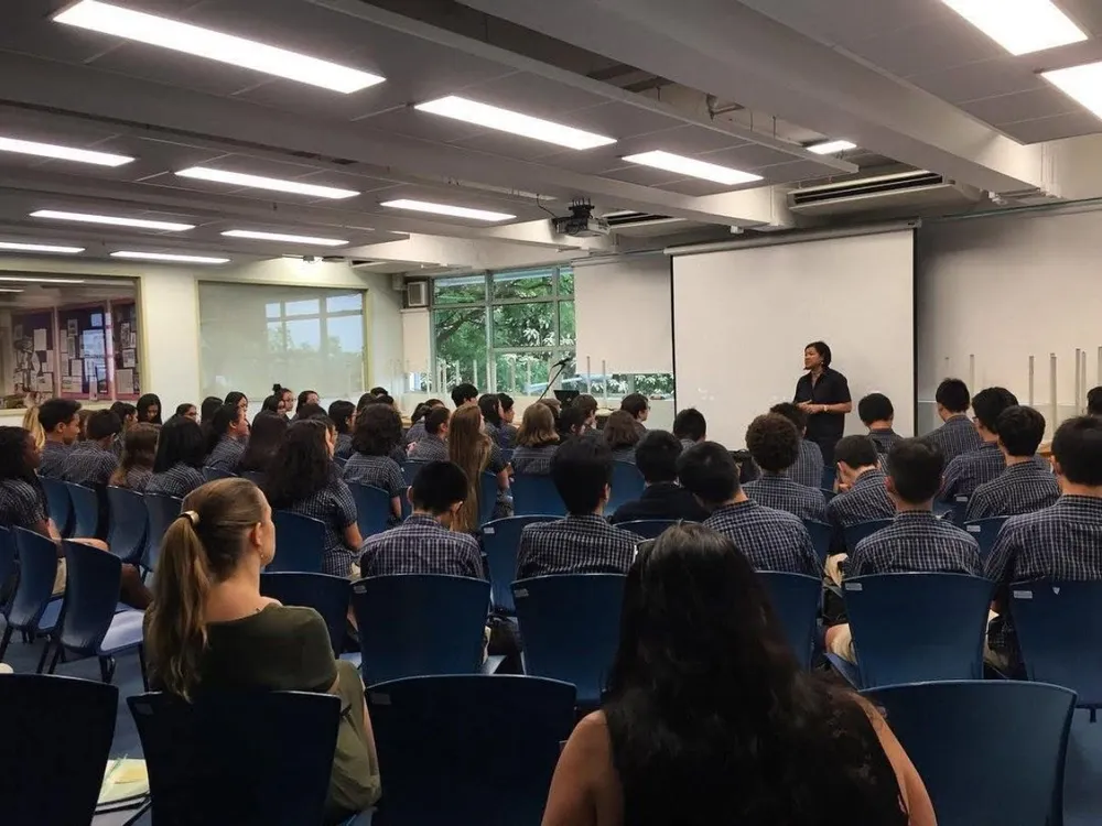 Speaking at the front of a bright hall to rows of students in school uniform, a projection screen behind.