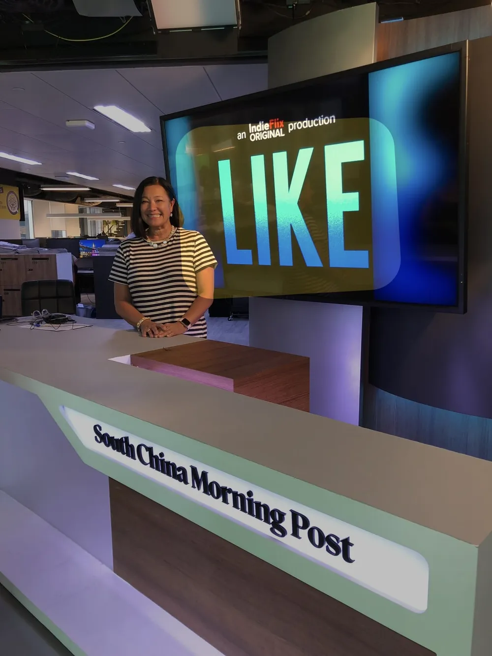 Scilla at the South China Morning Post studio desk, with LIKE on the big screen behind her.