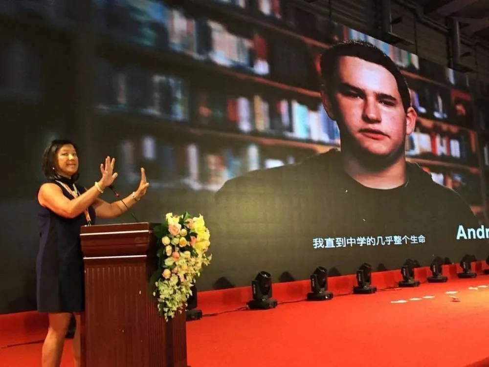 Speaking at a podium on a large stage with a red floor and flowers, beside a big screen showing a young man, subtitled in Chinese.