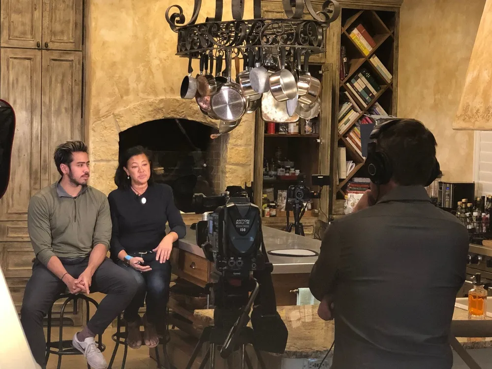 Ian and Scilla seated in a kitchen, facing a news camera and crew during an interview.