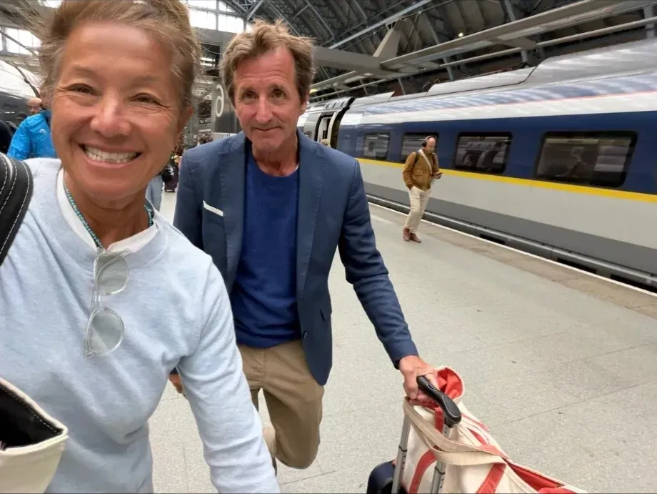 A smiling selfie of Scilla on a London train platform, her husband Eric behind her pulling a bag beside the train.
