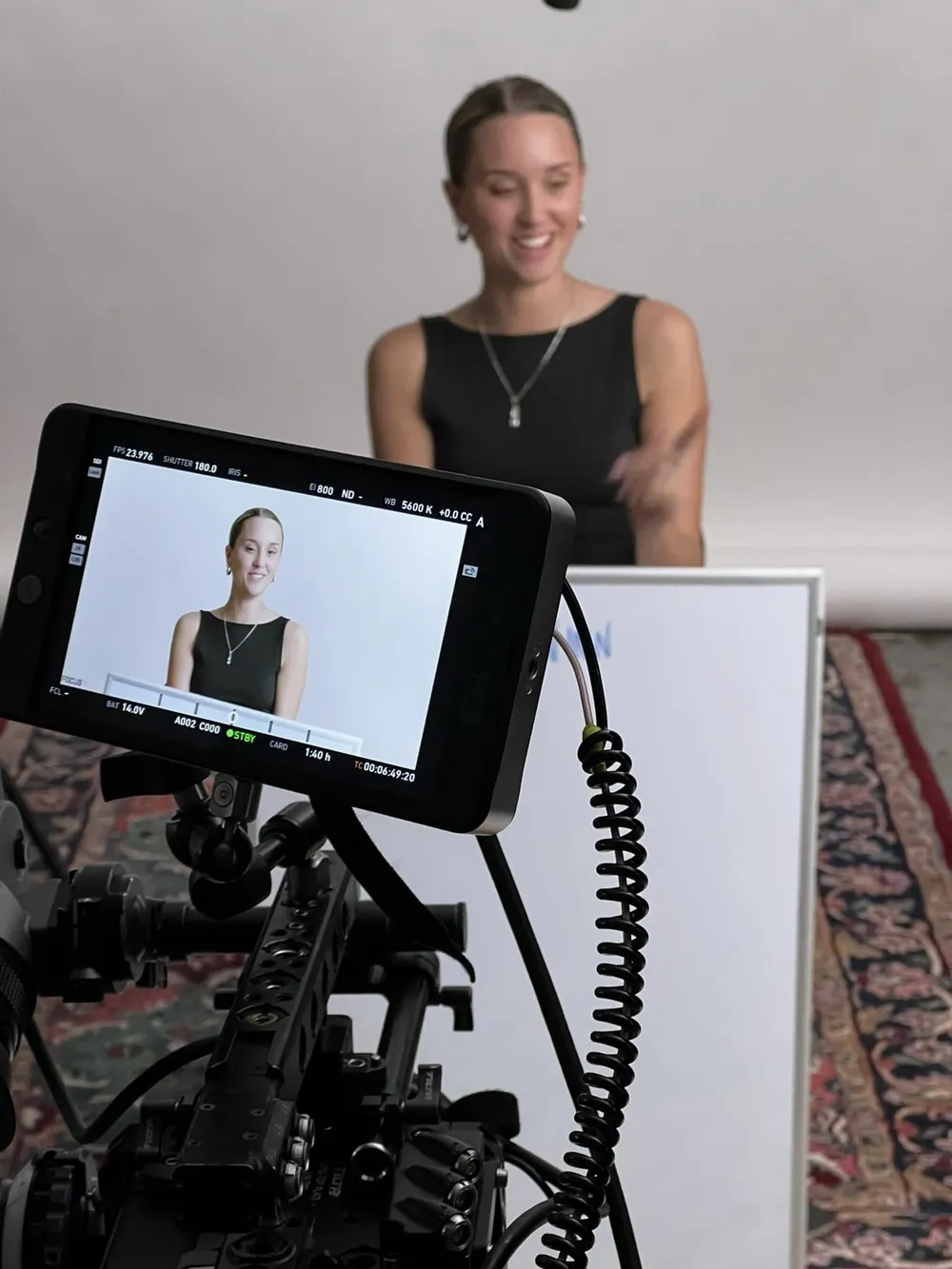 A young woman smiles in front of a white backdrop while a camera monitor in the foreground shows her framed for an interview.