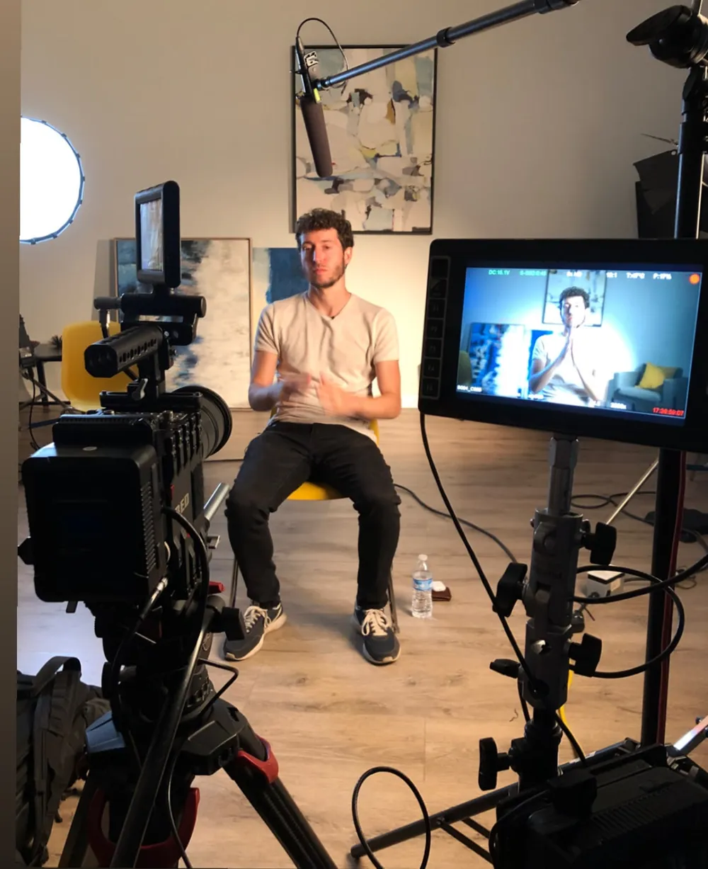 Max Stossel seated on a set being filmed, with cameras, lights and a monitor showing his interview.