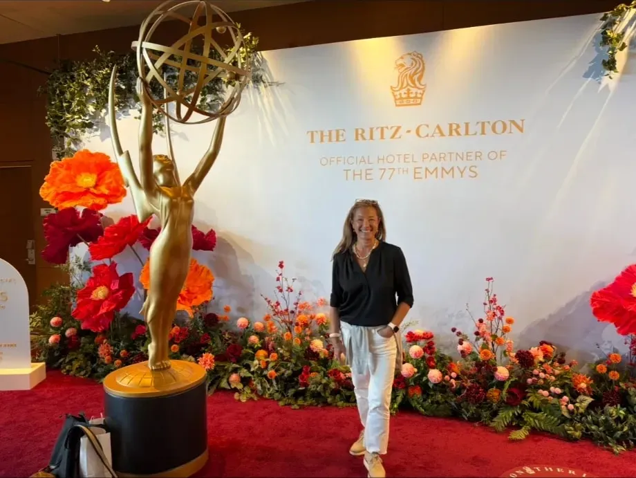 Scilla smiling beside a giant golden Emmy statue on a red carpet, in front of a backdrop for the 77th Emmys, framed by red and orange flowers.