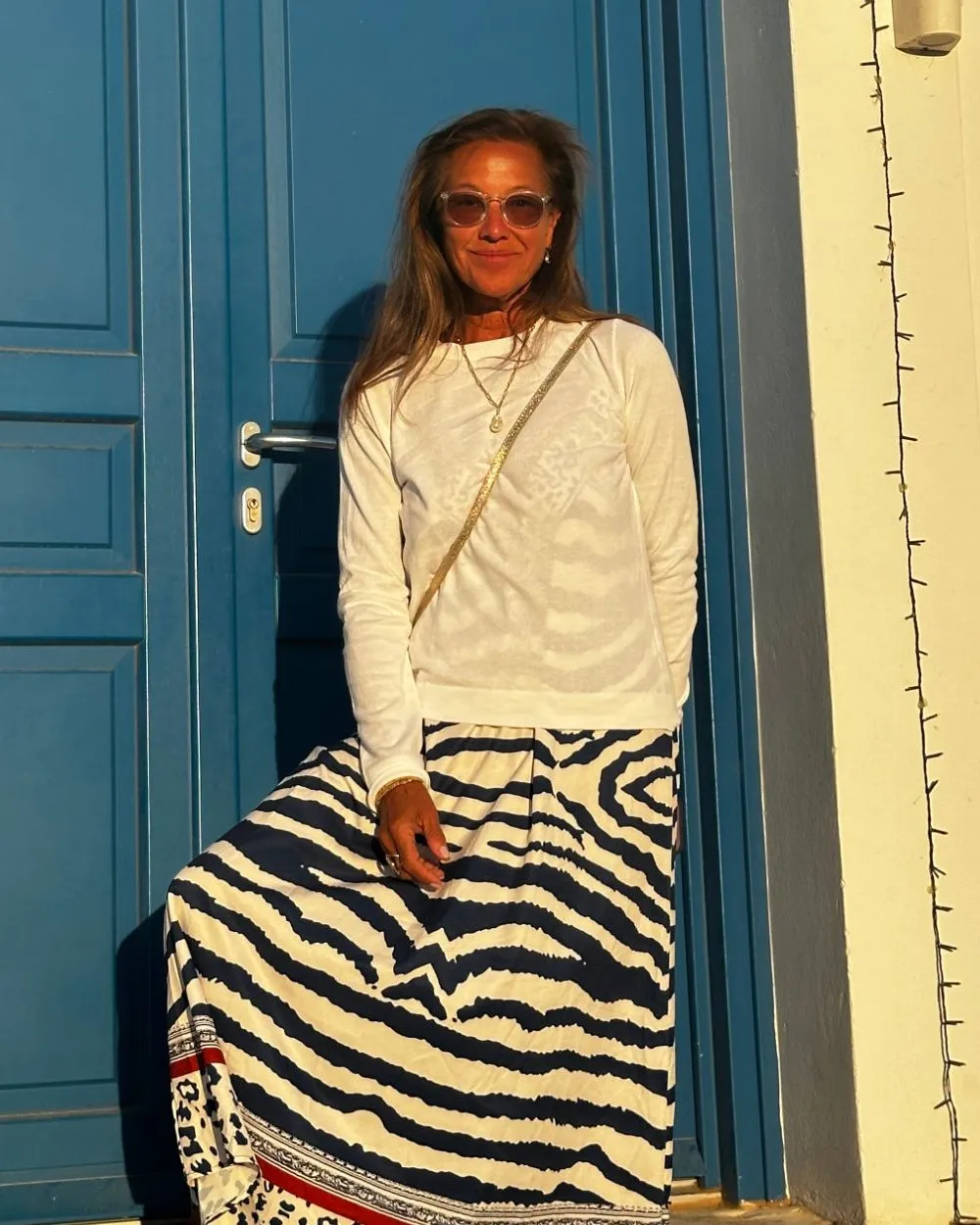 Scilla smiling in golden evening light, leaning against a blue door, in a white top and a long patterned skirt.