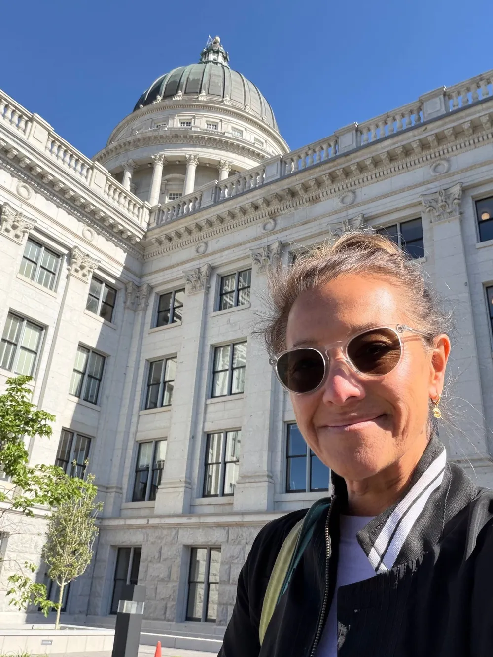 Scilla smiling in sunglasses in front of the Utah State Capitol, its dome against a clear blue sky.
