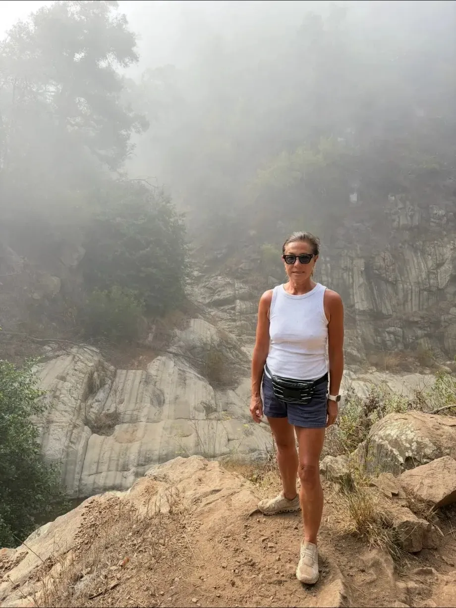 Scilla on a rocky trail in fog, in a white tank top and shorts, pale rock and trees behind her.