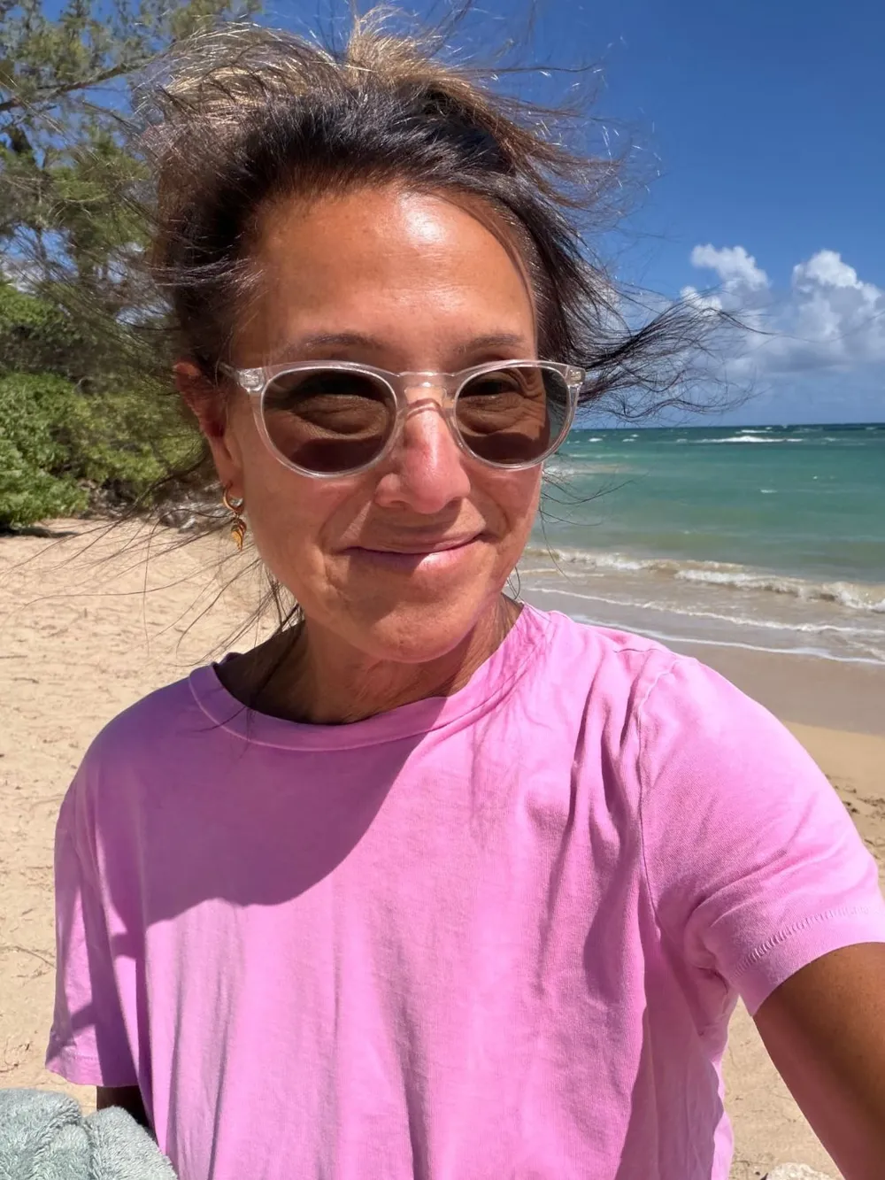 A windswept selfie of Scilla smiling on a Maui beach in a pink T-shirt and clear-framed sunglasses, turquoise sea behind her.