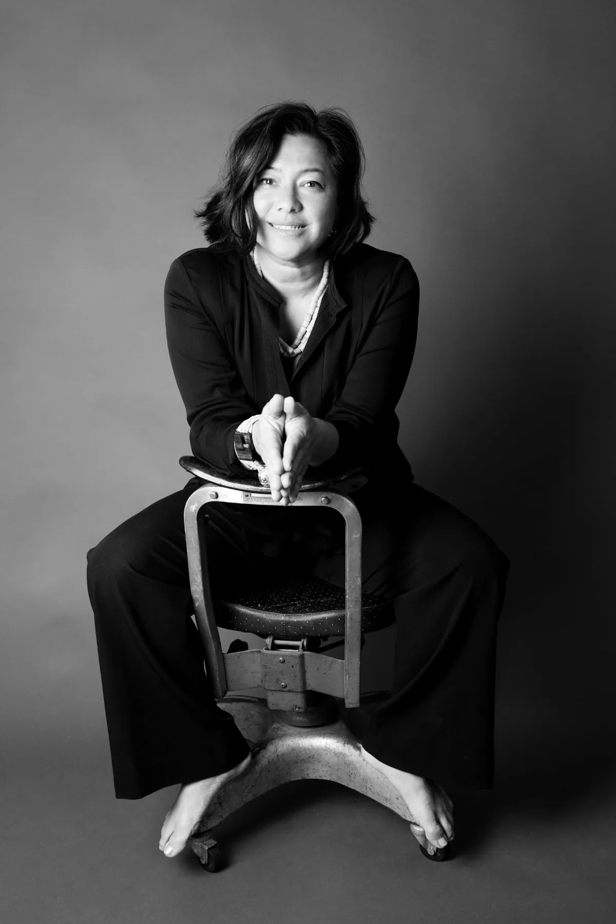Black-and-white studio portrait of Scilla, smiling, leaning forward on the back of a vintage metal chair, barefoot, in a black suit.
