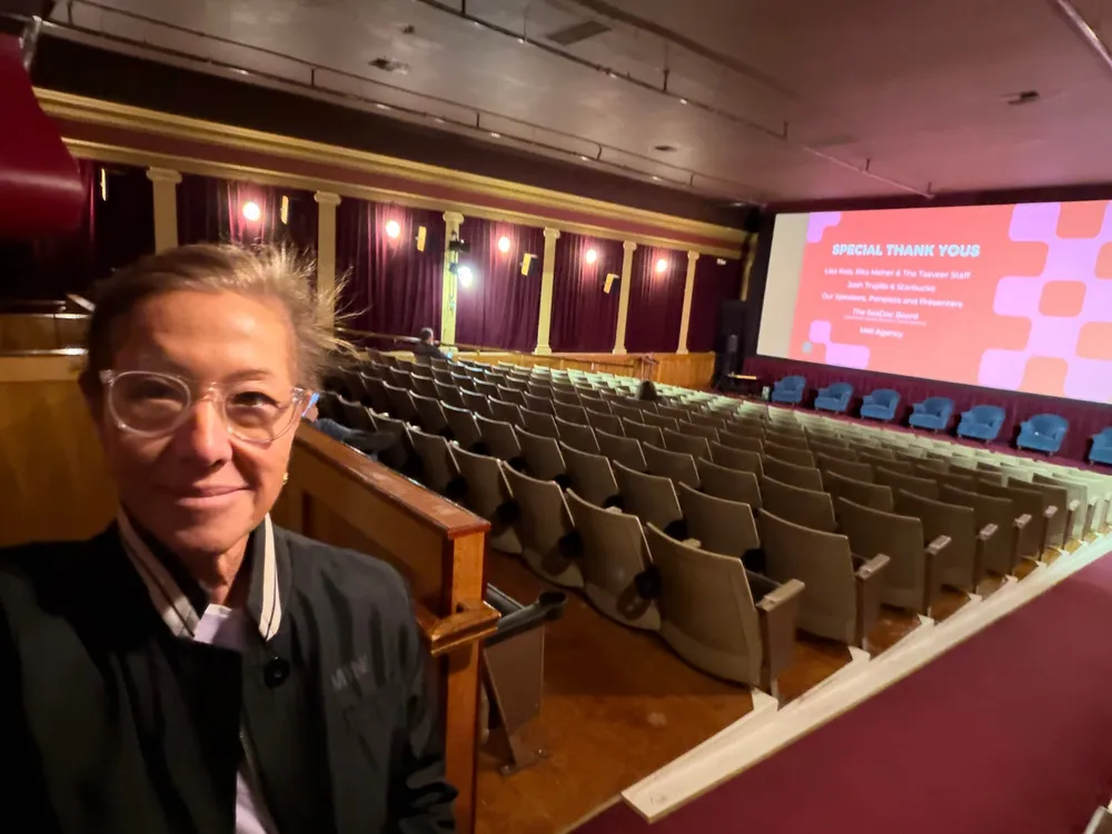 Scilla smiling in a cinema, rows of empty seats and a lit screen behind her.