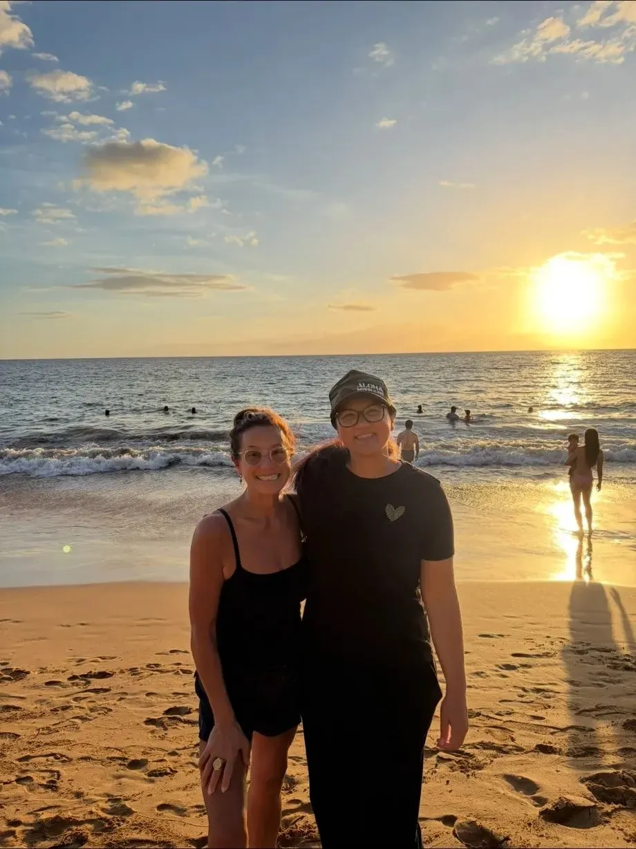 Scilla and Keala smiling together on a Maui beach at sunset, the sun low over the ocean behind them.