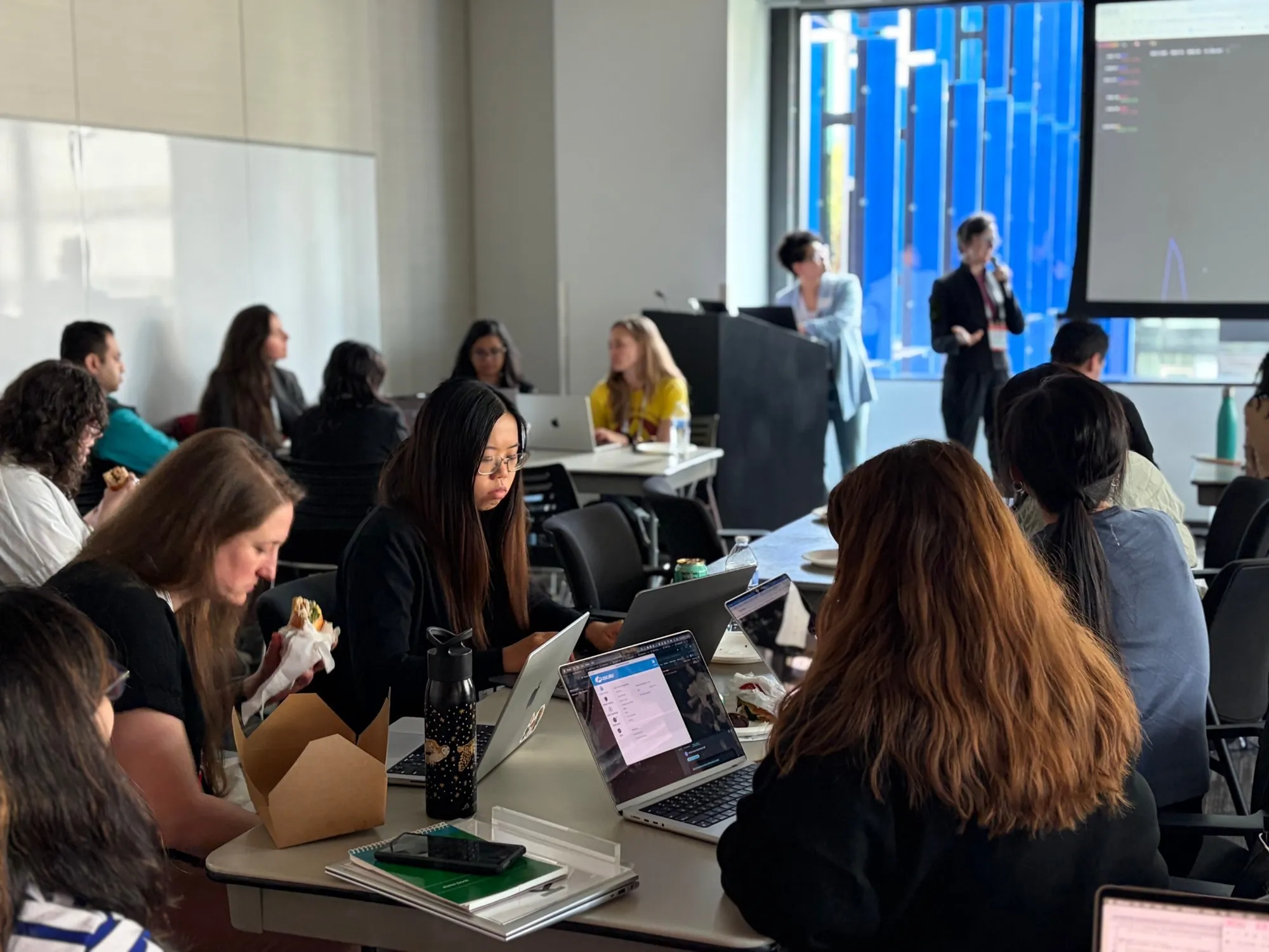 A bright room of women working at laptops around shared tables, with two presenters at the front beside a large screen.