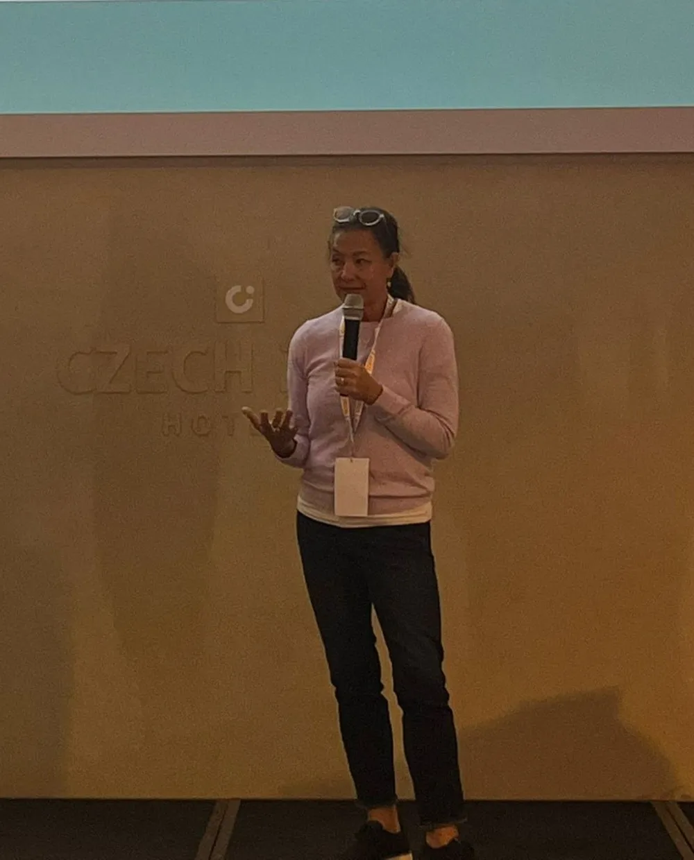 Scilla on stage in Prague, holding a microphone and gesturing as she speaks, in a lilac sweater with a conference badge.